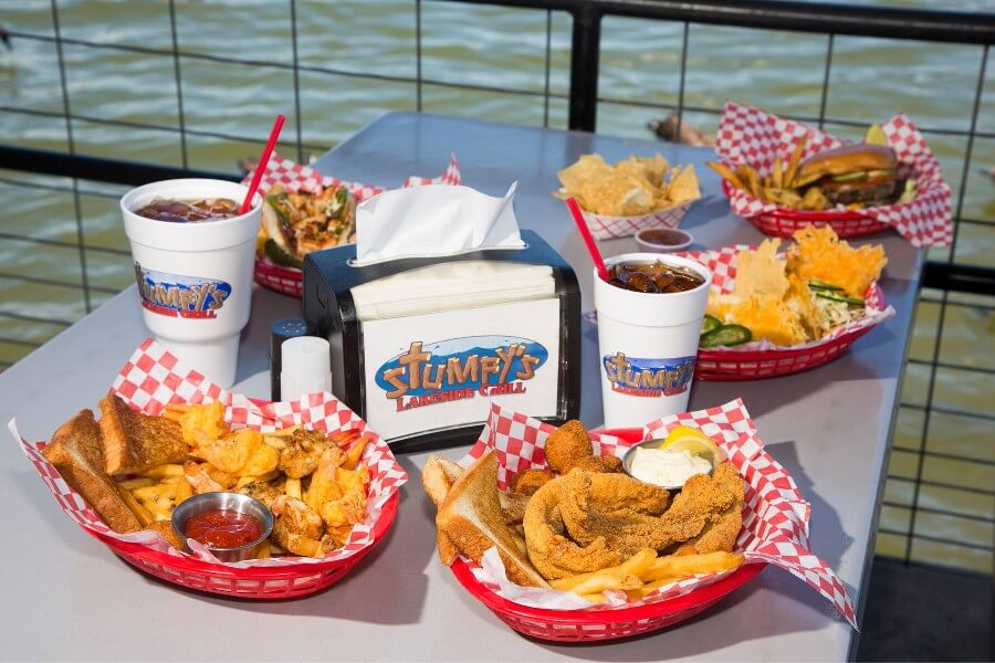 Variety of Stumpy’s Lakeside Grill meals in red baskets on a table with drinks and condiments, overlooking a water backdrop