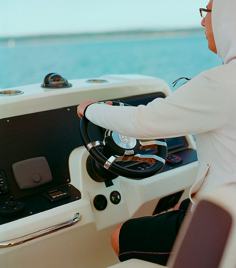 A man steering a boat, visible from behind, focusing on his hands on the wheel and the dashboard with navigational instruments