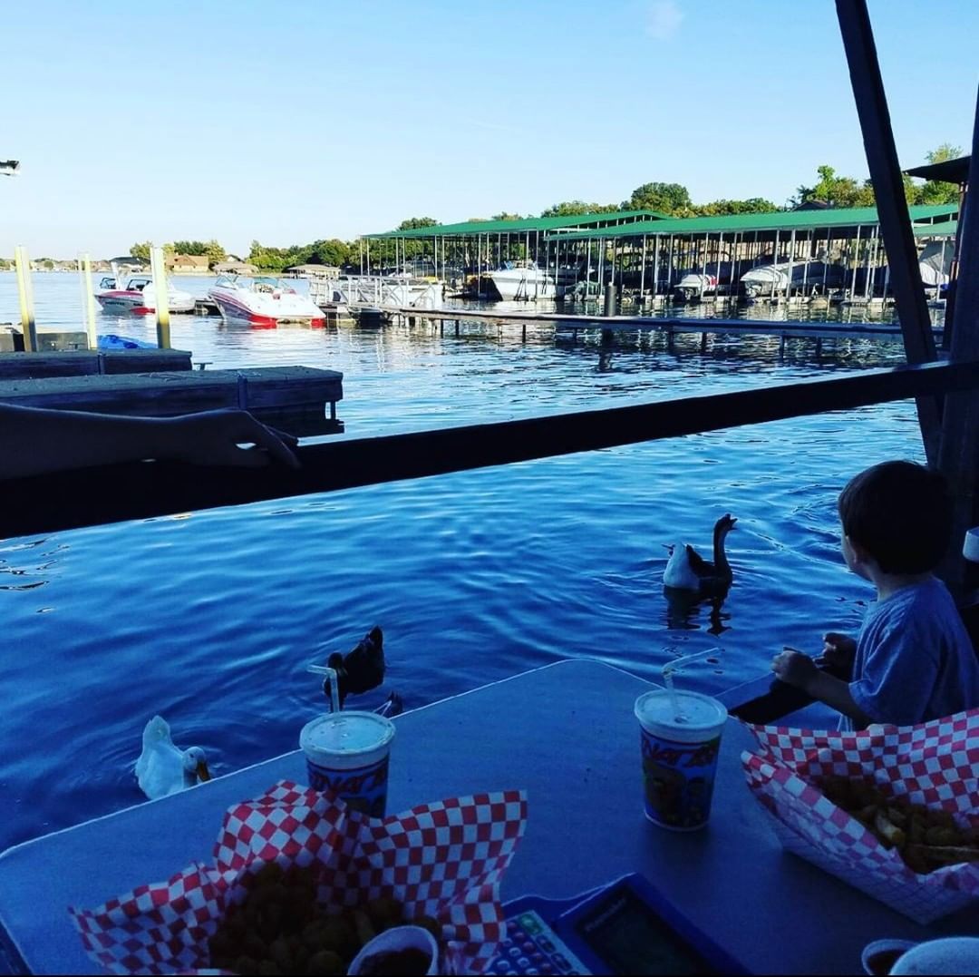 A child is at a waterfront dining table, feeding birds. Behind, Lake Granbury marina’s boat storage slips are visible under a blue sky